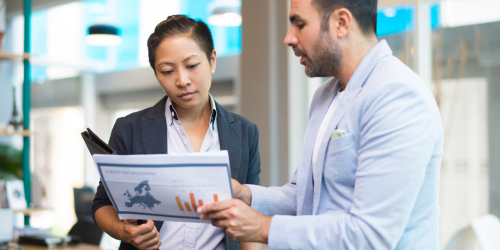 Brunette man with a short beard showing papers to a brunette woman with short hair in an office setting.