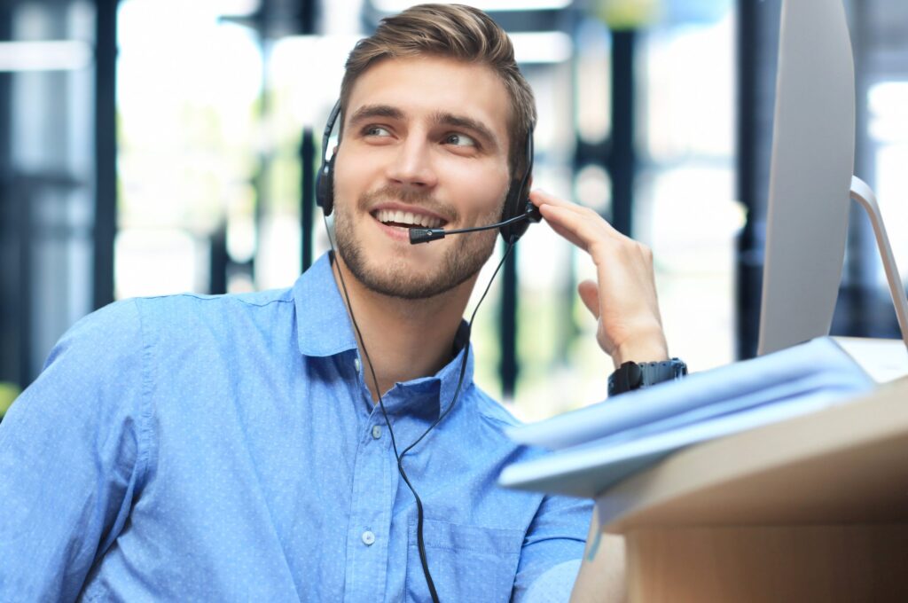 Smiling man in a blue shirt sitting at a computer wearing a wired communications headset.