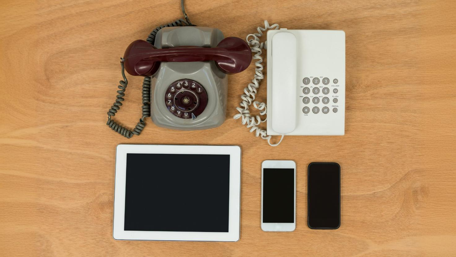 A flat lay of outdated and modern communication devices including a rotary phone, push-button landline phone, tablet, and two smartphones on a wooden surface. This image contrasts legacy phone systems with modern cloud-based phone systems, illustrating the evolution of business telecom solutions and the move toward scalable communication platforms like hosted PBX.
