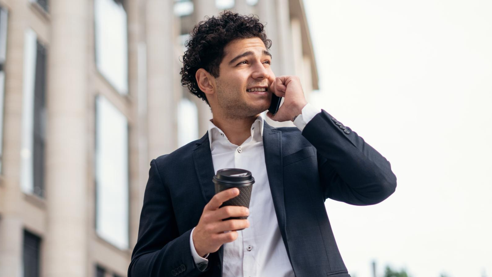 A smiling businessman in a suit holds a coffee cup while talking on a mobile phone outdoors, in front of a modern office building. This image reflects the flexibility and mobility enabled by hosted PBX phone systems and scalable, cloud-based business telecom solutions.