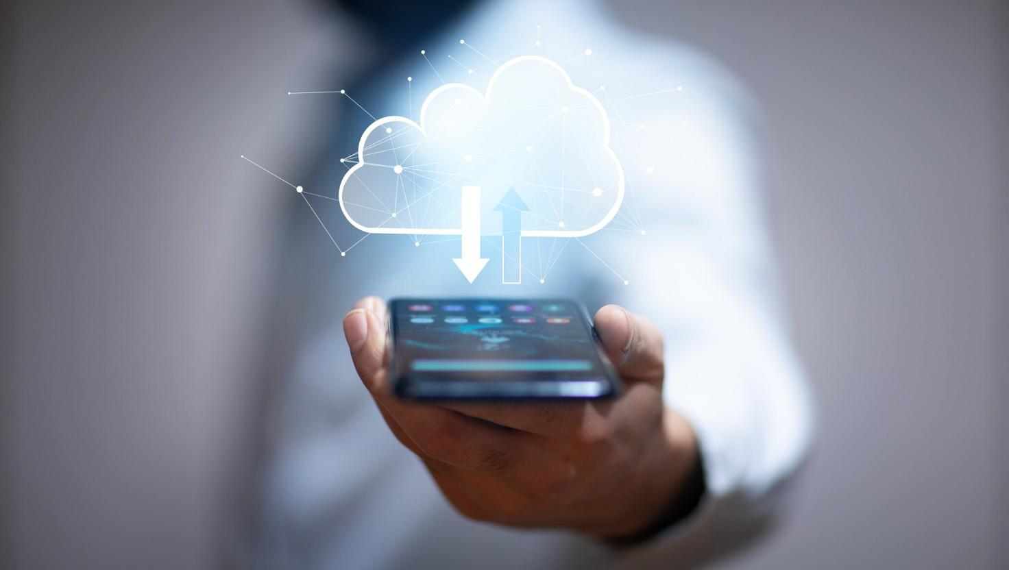 A person holds a smartphone in the foreground with a glowing cloud icon above it, featuring upload and download arrows. This image represents cloud-based phone systems and hosted PBX technology, emphasizing scalable communication and cloud-enabled business telecom solutions.