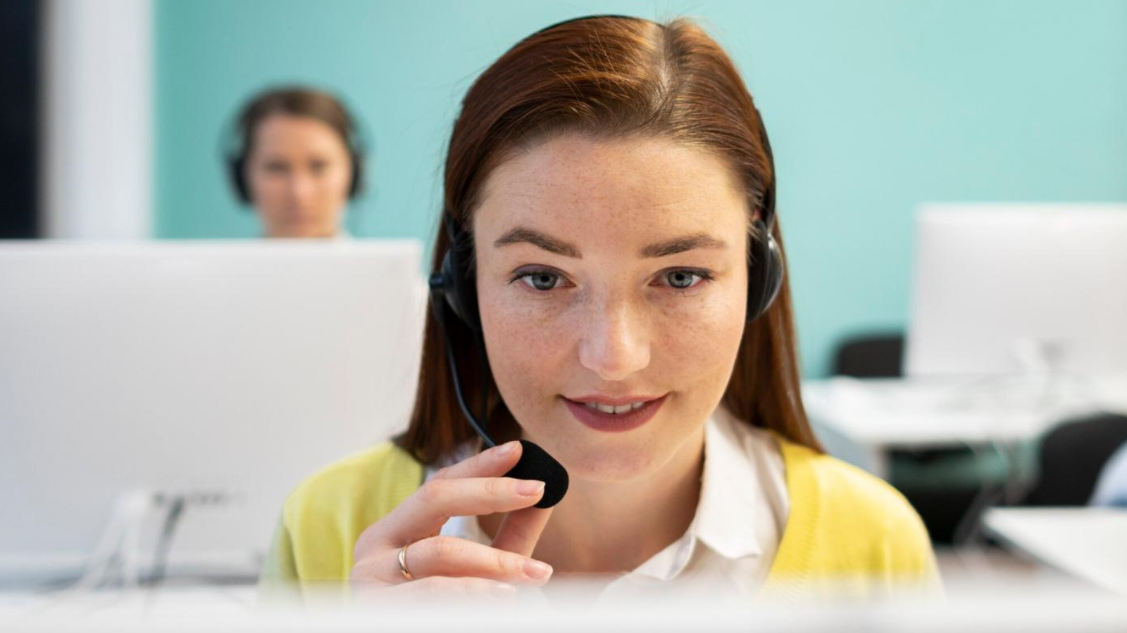 A close-up of a call center agent wearing a headset, speaking into a microphone with another agent blurred in the background. This image highlights real-time customer communication supported by business voice solutions and essential voice system features that enable effective use of telecom tools in service roles.