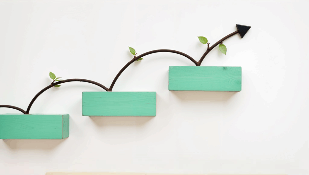 A conceptual image showing growth in scalable communication, featuring three ascending teal-colored wooden blocks mounted on a wall. A curved brown branch with small green leaves and a black arrow tip grows upward across the blocks, symbolizing progressive phone system upgrades and the evolution of modern telecom tools in office phone systems.
