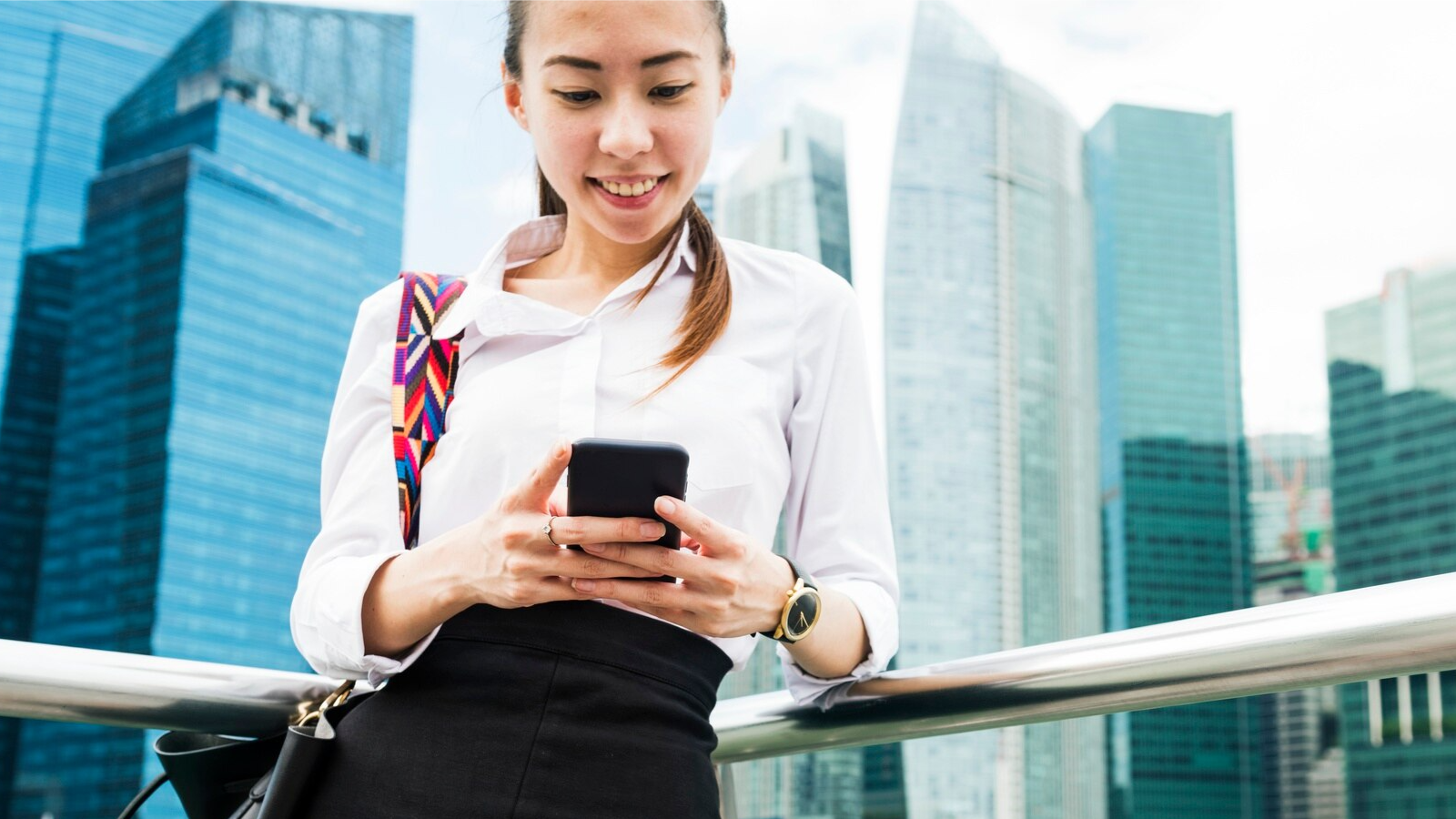A professional woman in business attire smiles while using her smartphone in an urban setting with modern skyscrapers. The scene emphasizes the mobility and flexibility of modern telecom tools and scalable communication, representing how office phone systems and phone system upgrades empower remote and urban professionals alike.