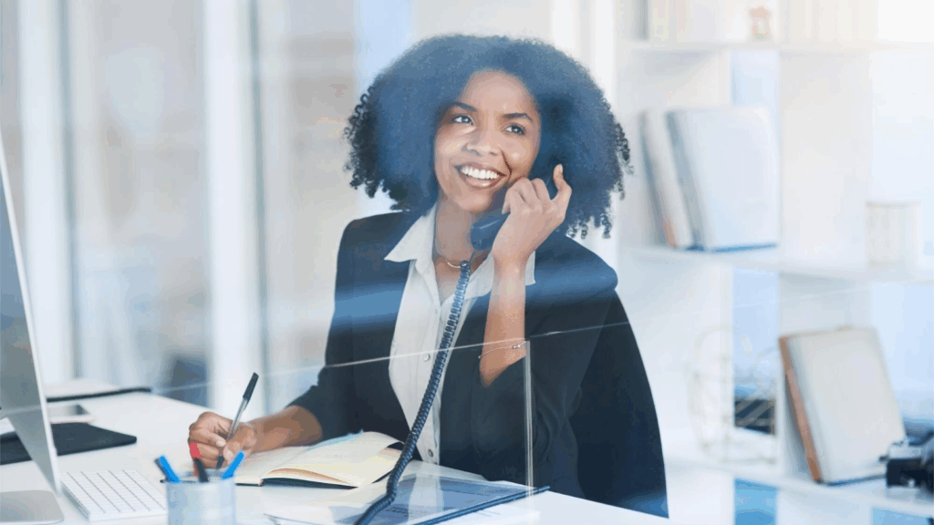 A smiling professional woman speaks on a corded office phone while taking notes at her desk in a bright, modern office. She appears engaged and productive, representing how office phone systems and modern telecom tools support efficient communication. The setting highlights the value of phone system upgrades and scalable communication solutions for today’s workplace.