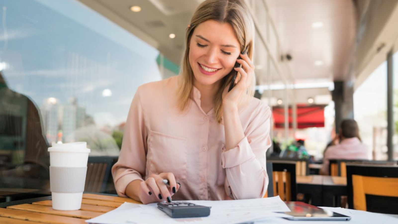 Woman at a café smiling during a phone call while using a calculator, highlighting cloud services that improve cost & operational efficiency.